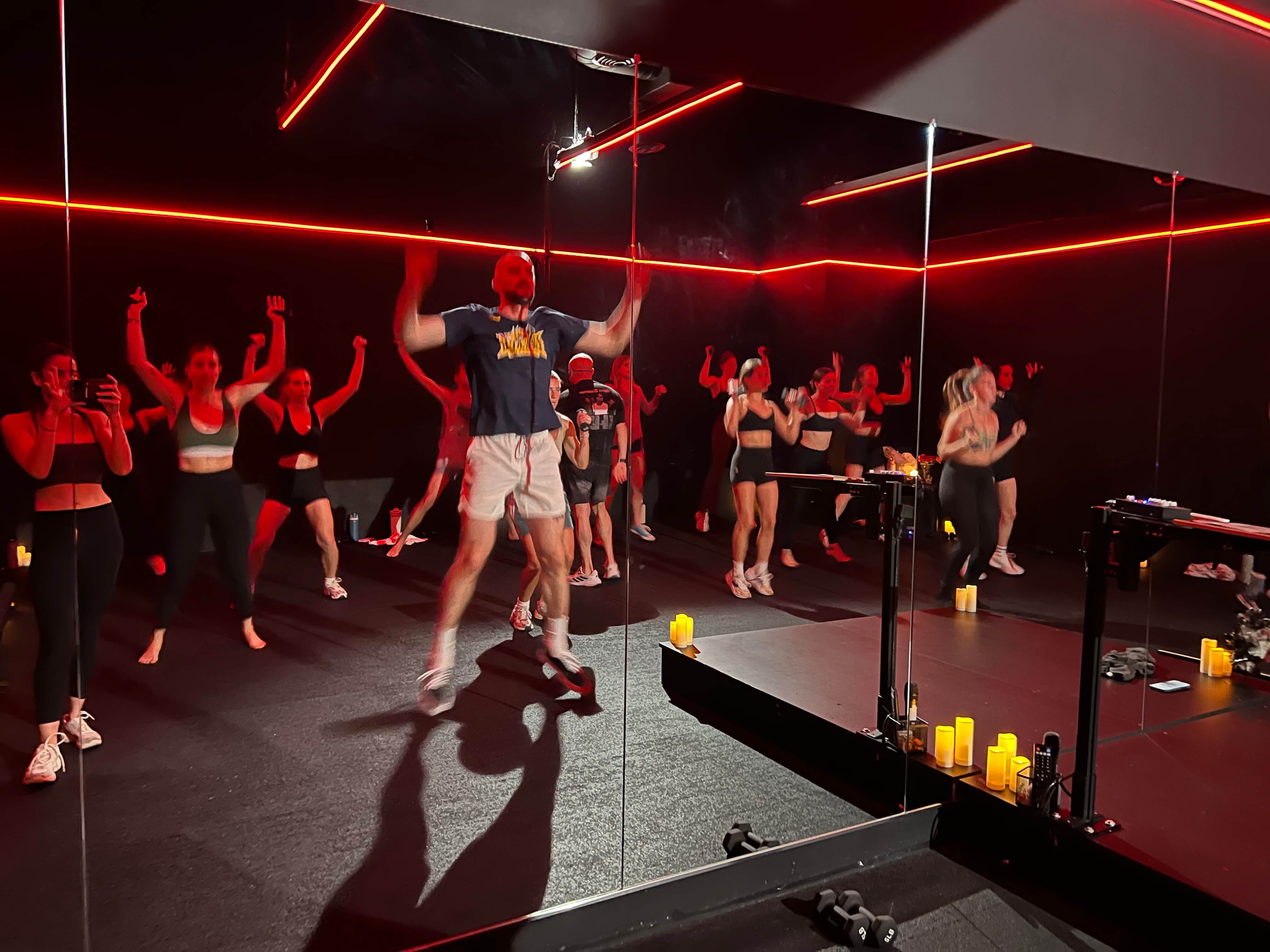 Group fitness class with instructor performing jumping exercise under red neon lights and mirrored walls